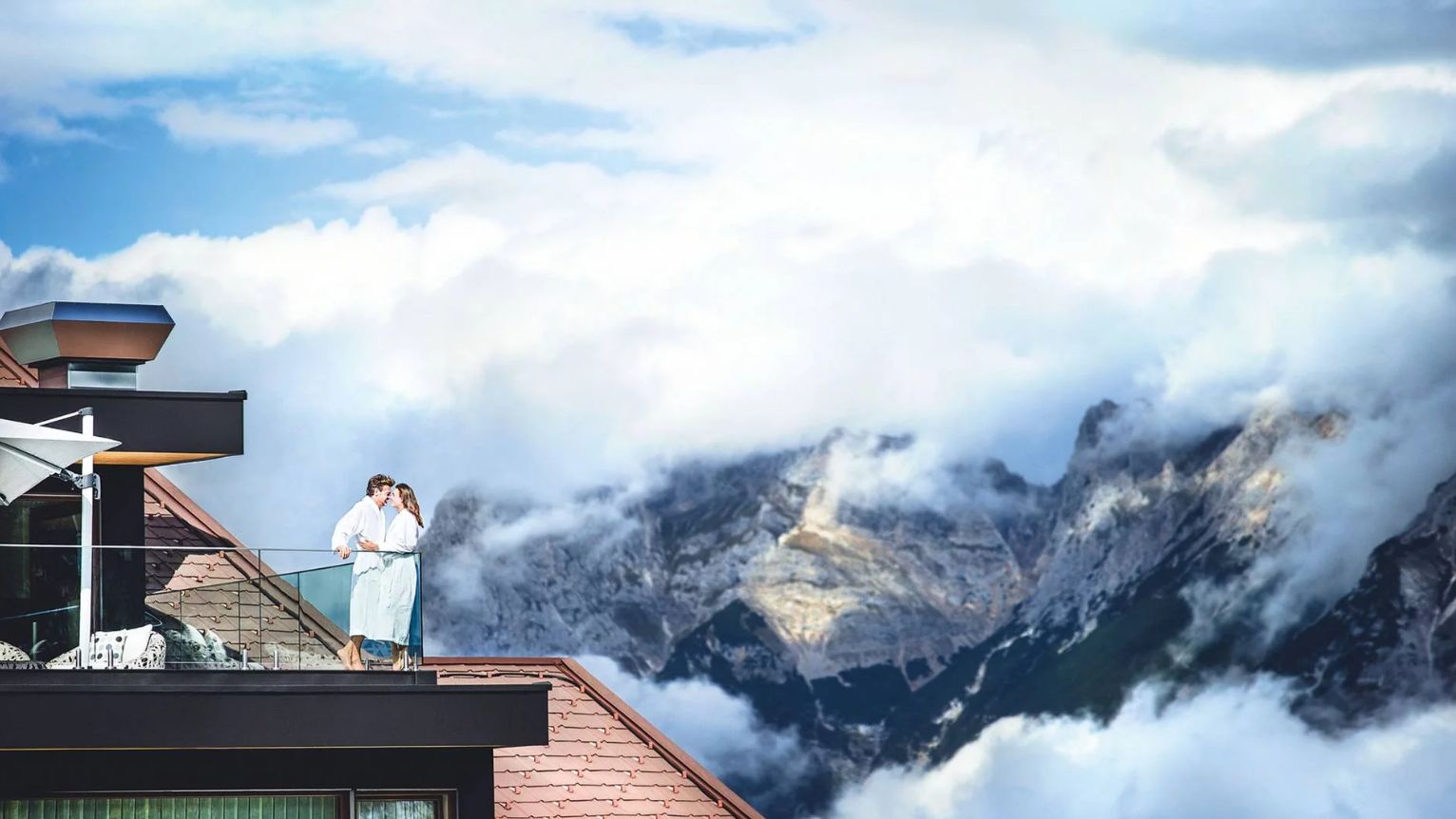 Ein Paar in Bademänteln steht auf einem Hoteldachbalkon und blickt auf eine beeindruckende Bergkulisse, teils von Wolken umhüllt – ein Moment der Ruhe und Zweisamkeit in den Alpen | © 2025 Hotel Klosterbräu - Seefeld in Tirol