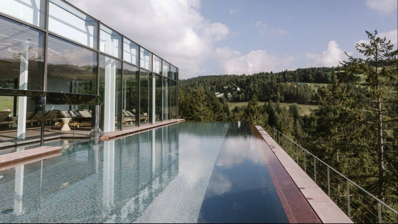Infinity-Pool eines modernen Wellnesshotels mit Glasfassade, umgeben von Wald und sanfter Hügellandschaft | © Hotel Saltus