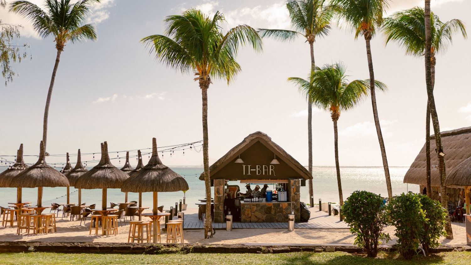 Strandbar mit dem Namen „Ti-Bar“ unter Palmen an einem tropischen Strand, mit Strohdach, Steinsockel und Barhockern. Links davon stehen mehrere mit Strohdächern überdachte Sitzbereiche mit Tischen und Stühlen direkt am Meer. Im Hintergrund ruhiges türkisfarbenes Wasser und ein heller Himmel mit wenigen Wolken | © Sun Hotels & Resorts GmbH