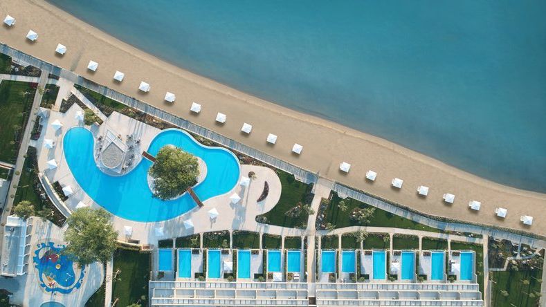 Overview auf das Ikos Dassia mit mehreren Pools, gepflegten Gartenwegen und einem angrenzenden Sandstrand mit weißen Sonnenschirmen am türkisblauen Meer | © Ikos