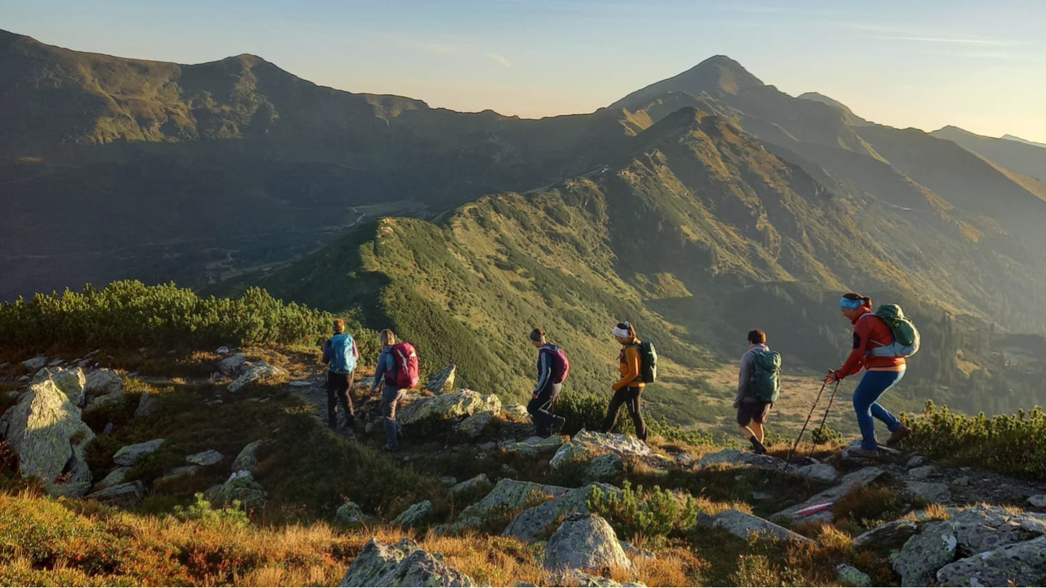 Eine Wandergruppe ist auf einem felsigen Wanderweg unterwegs, während morgendliche Sonnenstrahlen die Berglandschaft in warmes Licht tauchen.